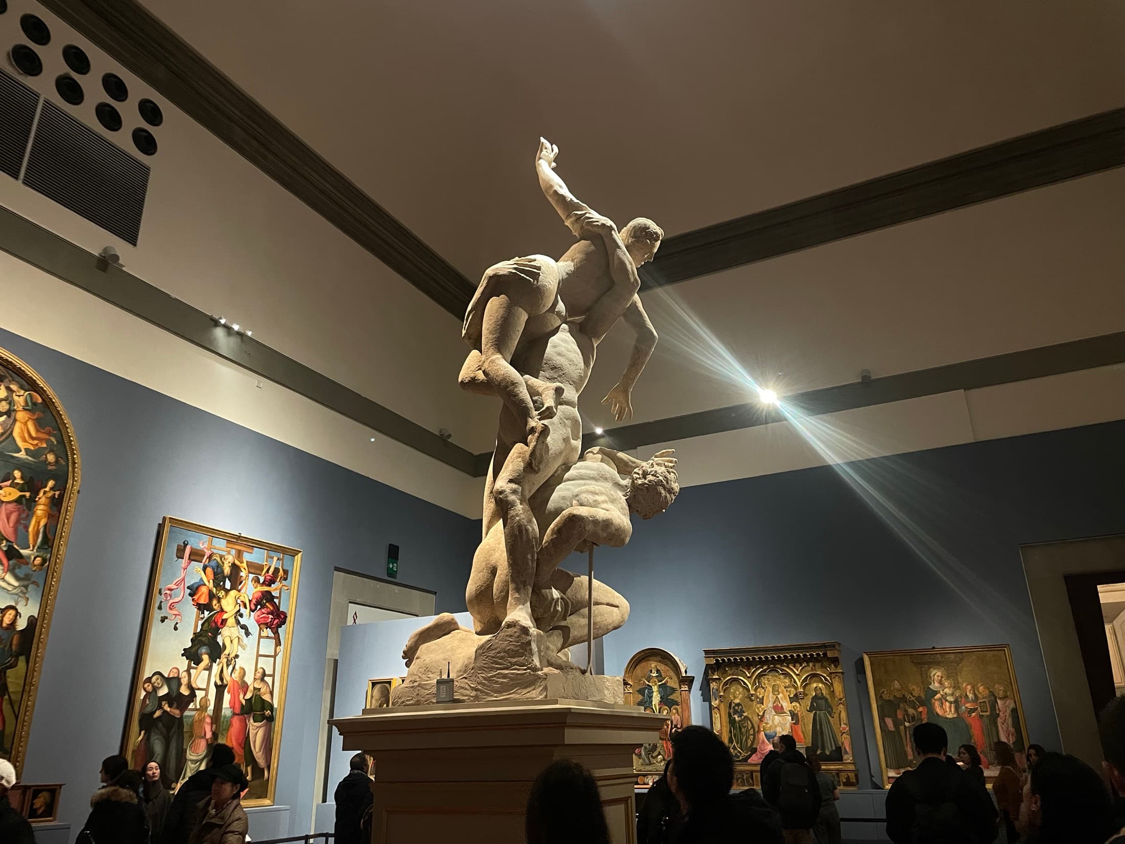 A marble sculpture group lit by a shaft of light in a museum hall, gilt-framed paintings on the walls behind