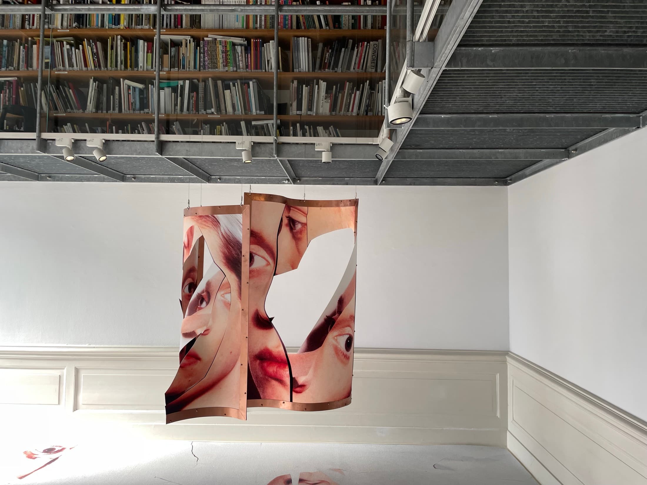 Large printed panels of cropped faces hung in a room below a wall of bookshelves