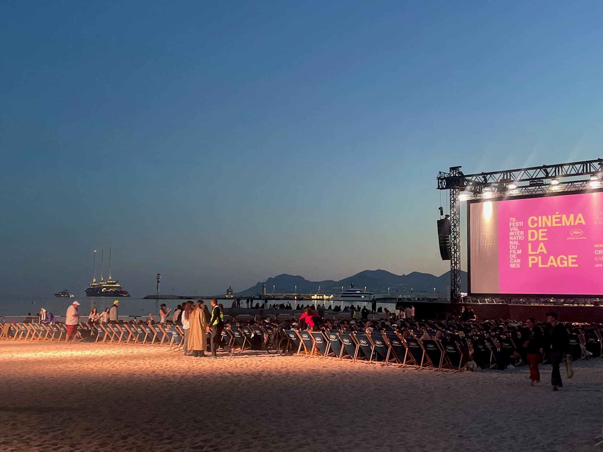 Rows of chairs on a beach at dusk in front of an open-air cinema screen