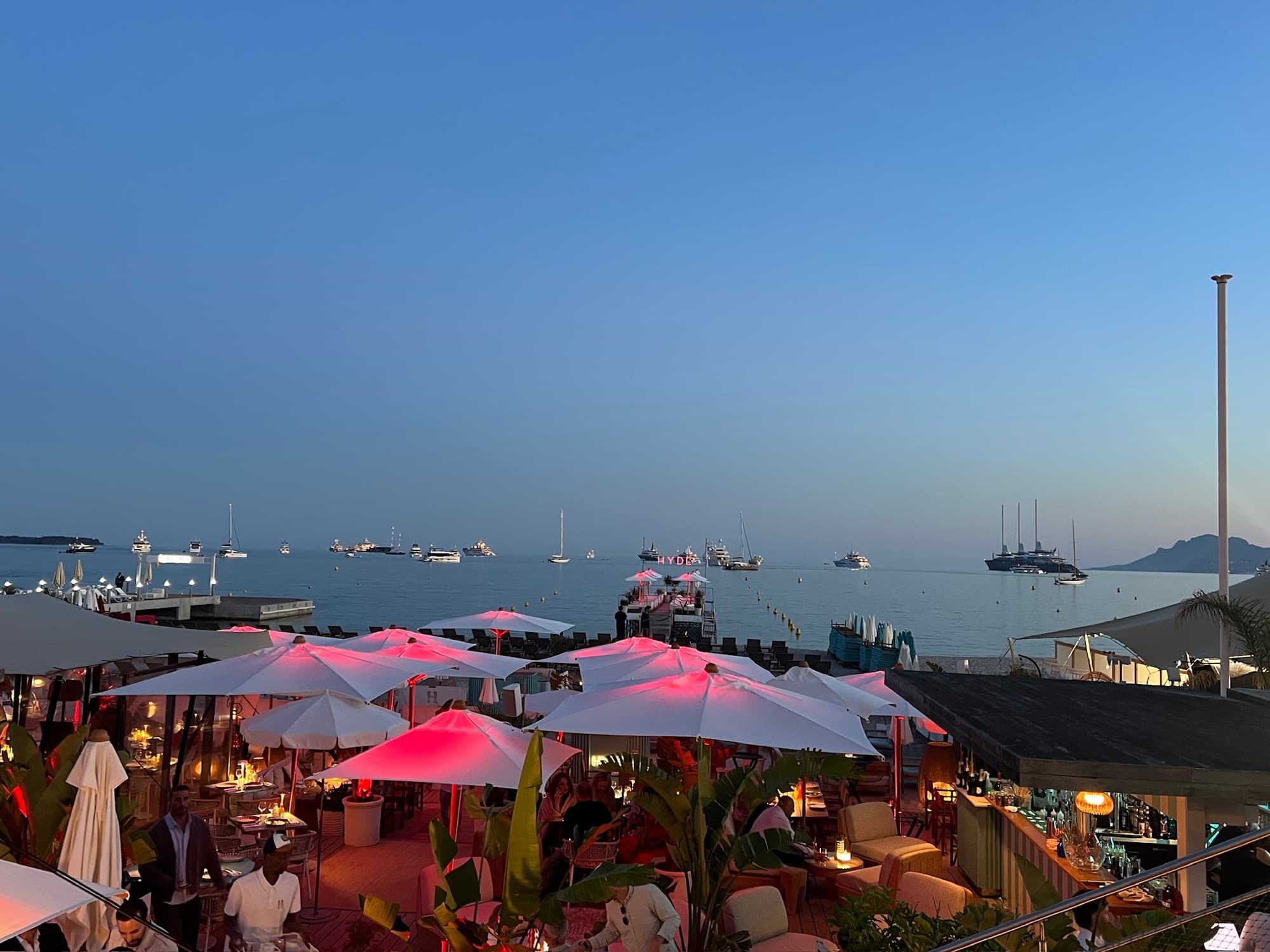 White parasols over a seaside terrace at dusk, boats on the water behind