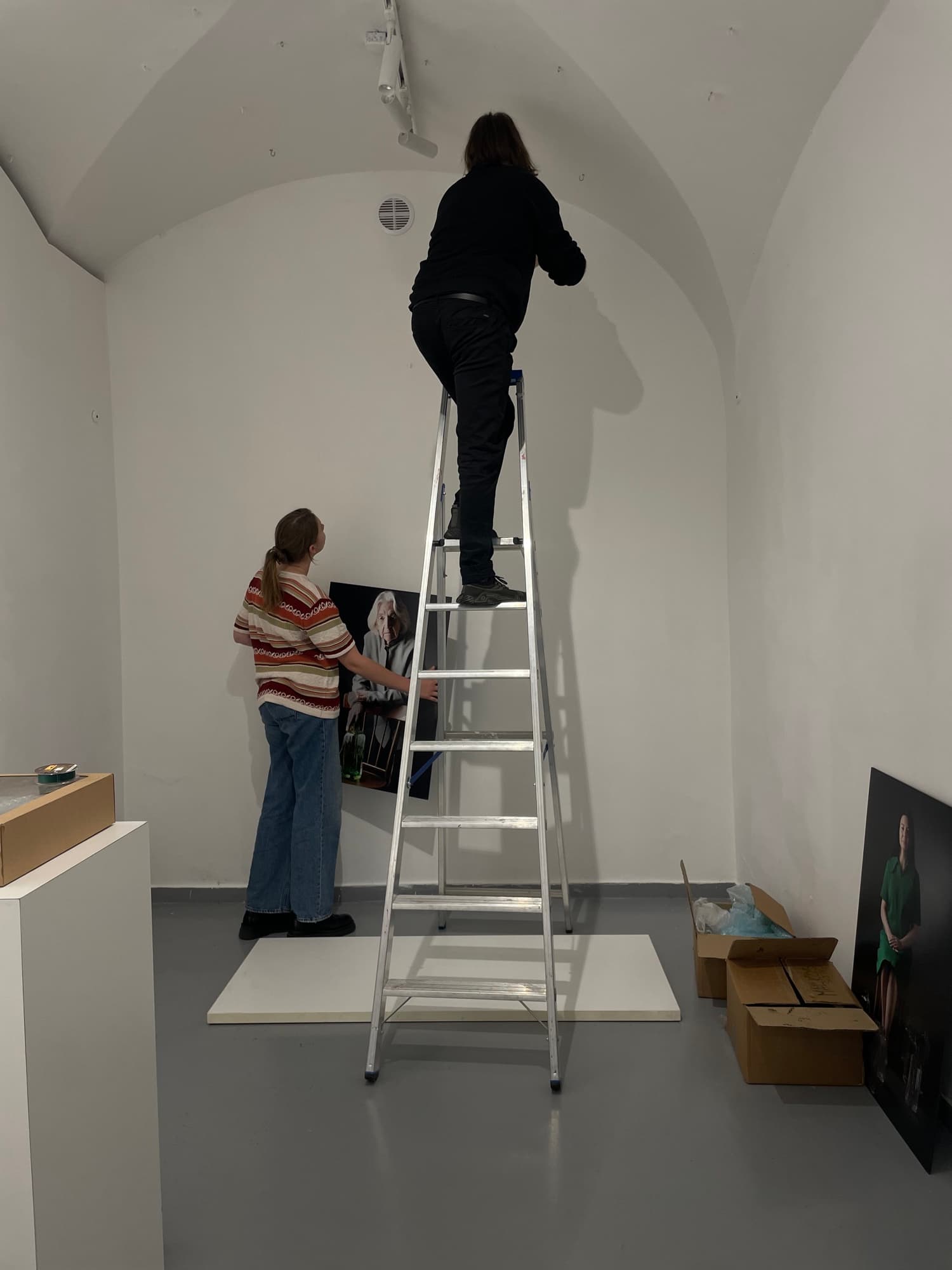 Two people hanging a framed photograph on a white gallery wall, one up a stepladder
