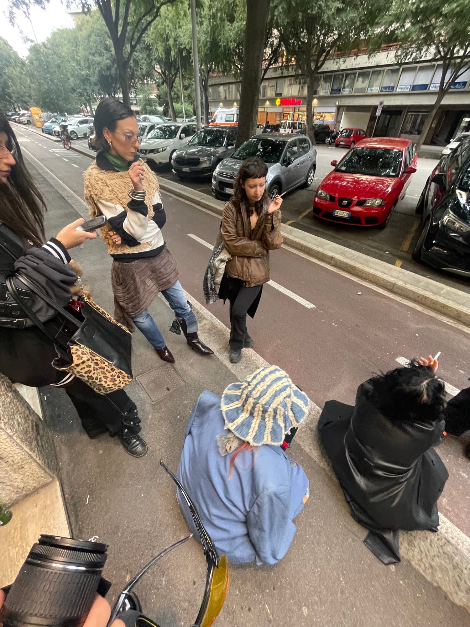 Four people in coats and boots crossing a street between parked cars, one carrying a leopard-print bag, another lighting a cigarette