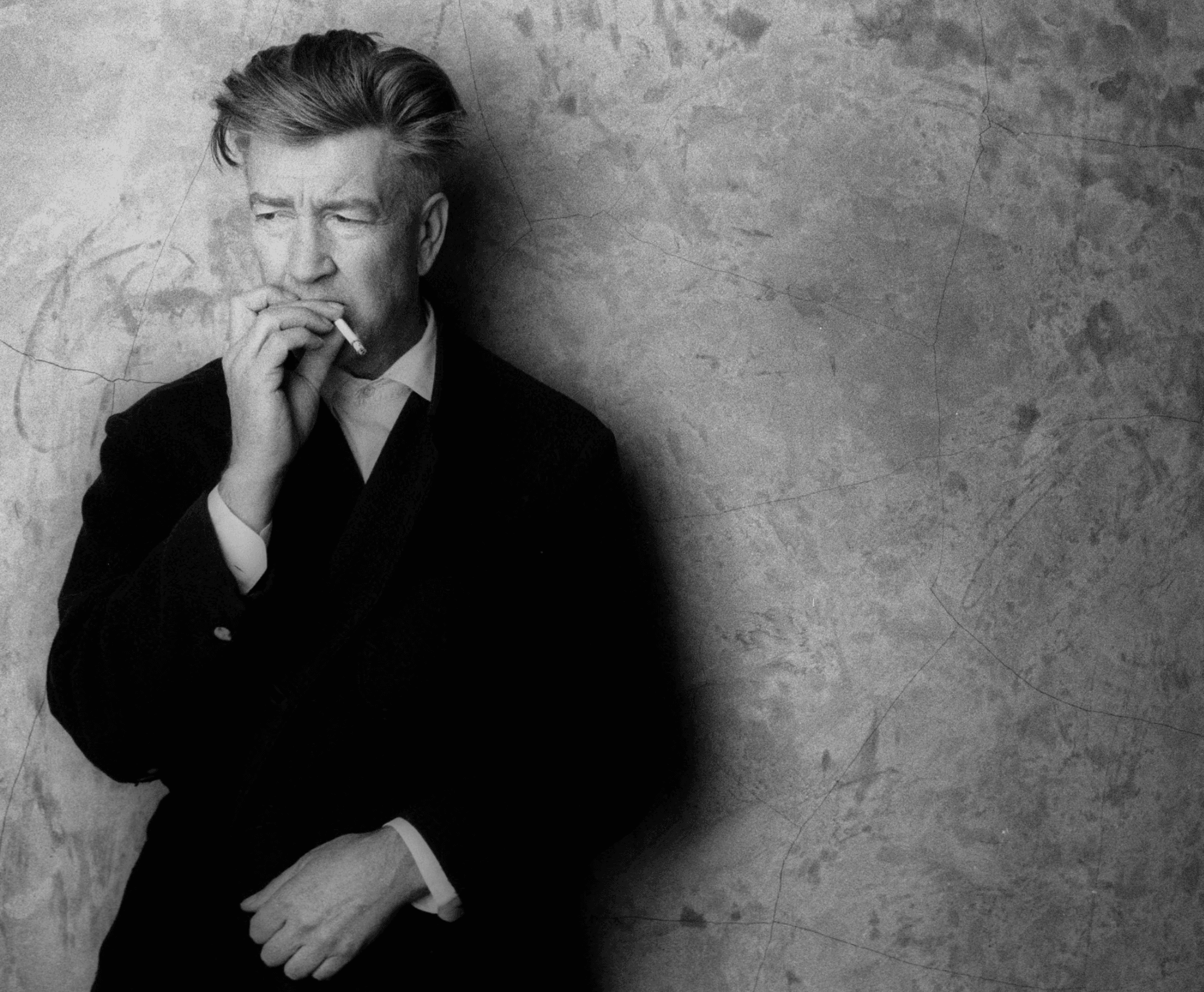 David Lynch, in a dark coat, leans against a cracked plaster wall smoking a cigarette, in a black and white portrait.