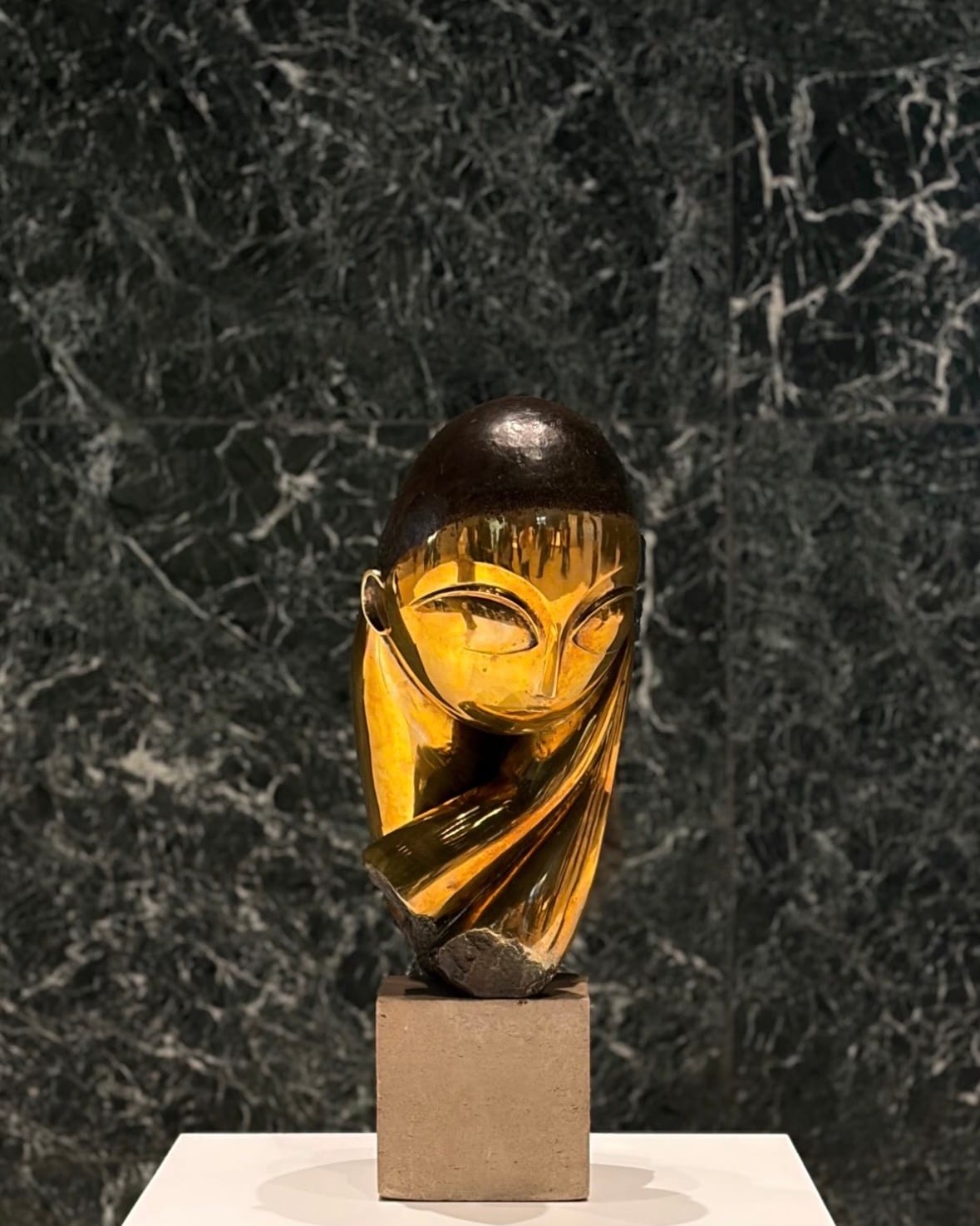 A polished bronze head by Constantin Brancusi on a stone plinth, lit against a dark green marble wall.