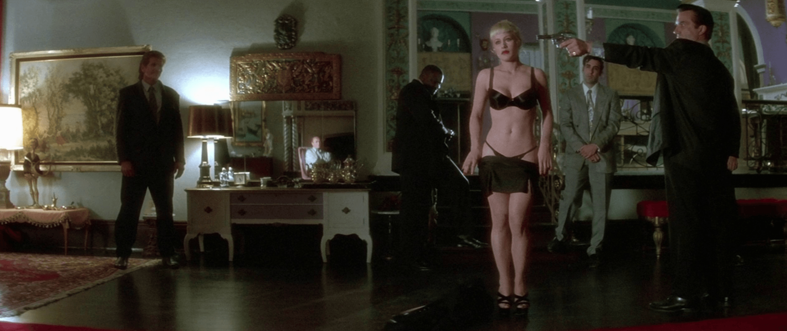 A woman in black lingerie stands in a dim, ornate living room while a man in a dark suit aims a handgun at her and several other men look on, a still from the film Lost Highway.
