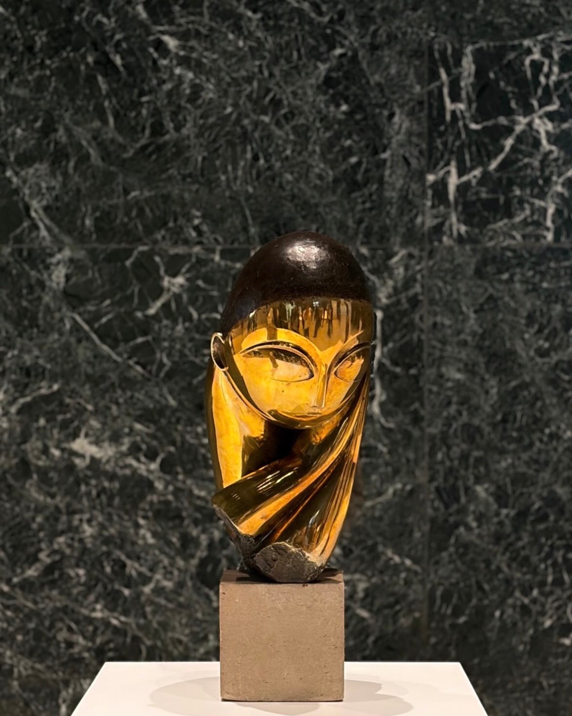 A polished bronze head by Constantin Brancusi on a stone plinth, lit against a dark green marble wall.