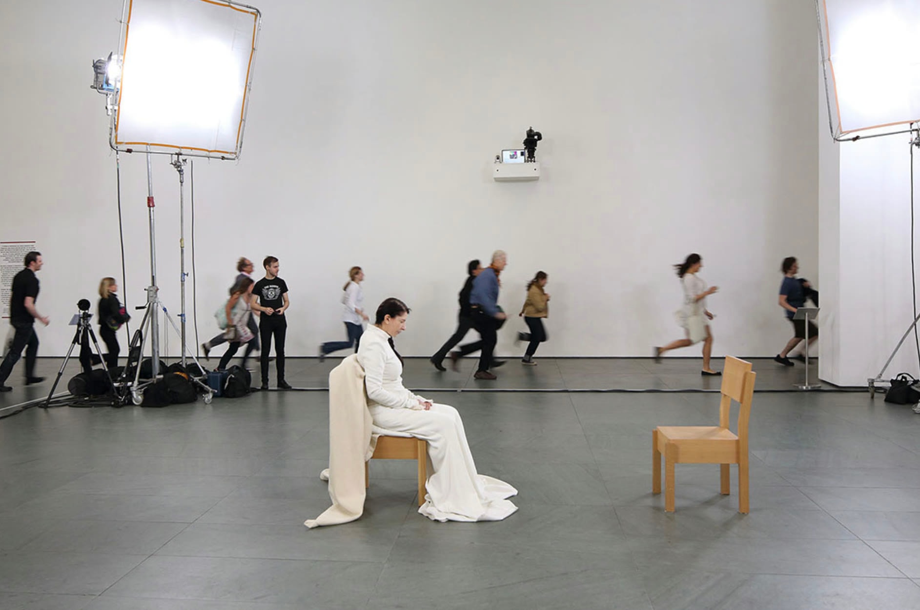 Marina Abramović seated in a white gown at MoMA, an empty wooden chair opposite her, film lights either side and visitors blurred with movement behind.