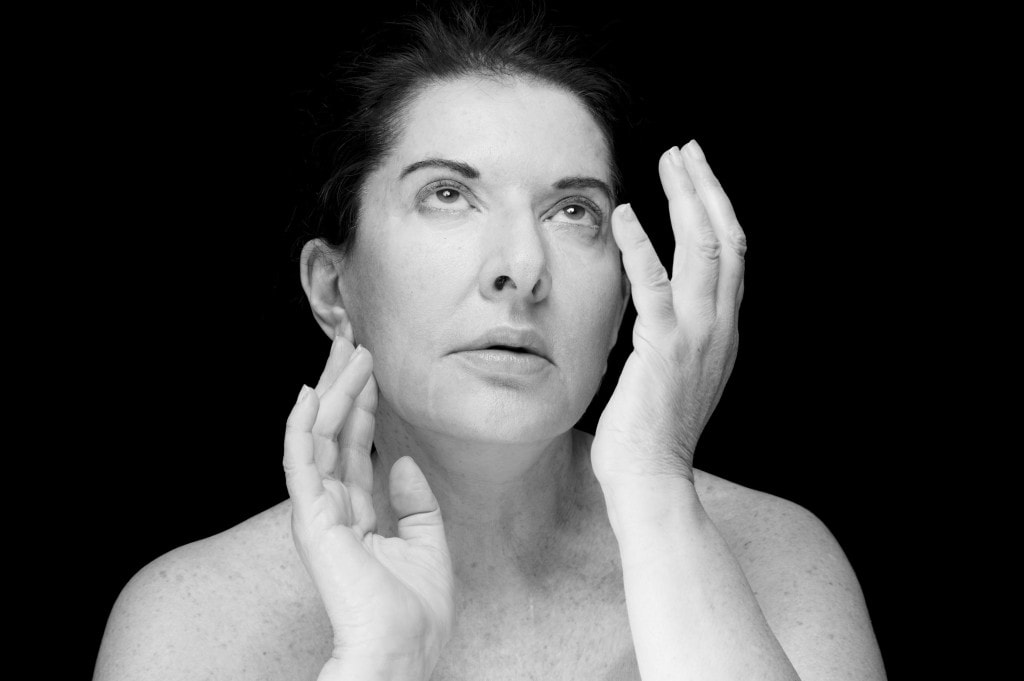 Marina Abramović photographed against black, both hands raised to her face, looking upward.