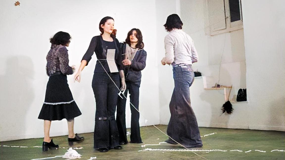 A performance in a bare room: a woman stands with cords stretched from her body while three other people move around her.