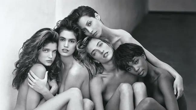 Five models photographed in black and white, leaning into one another in a close group against a plain wall.