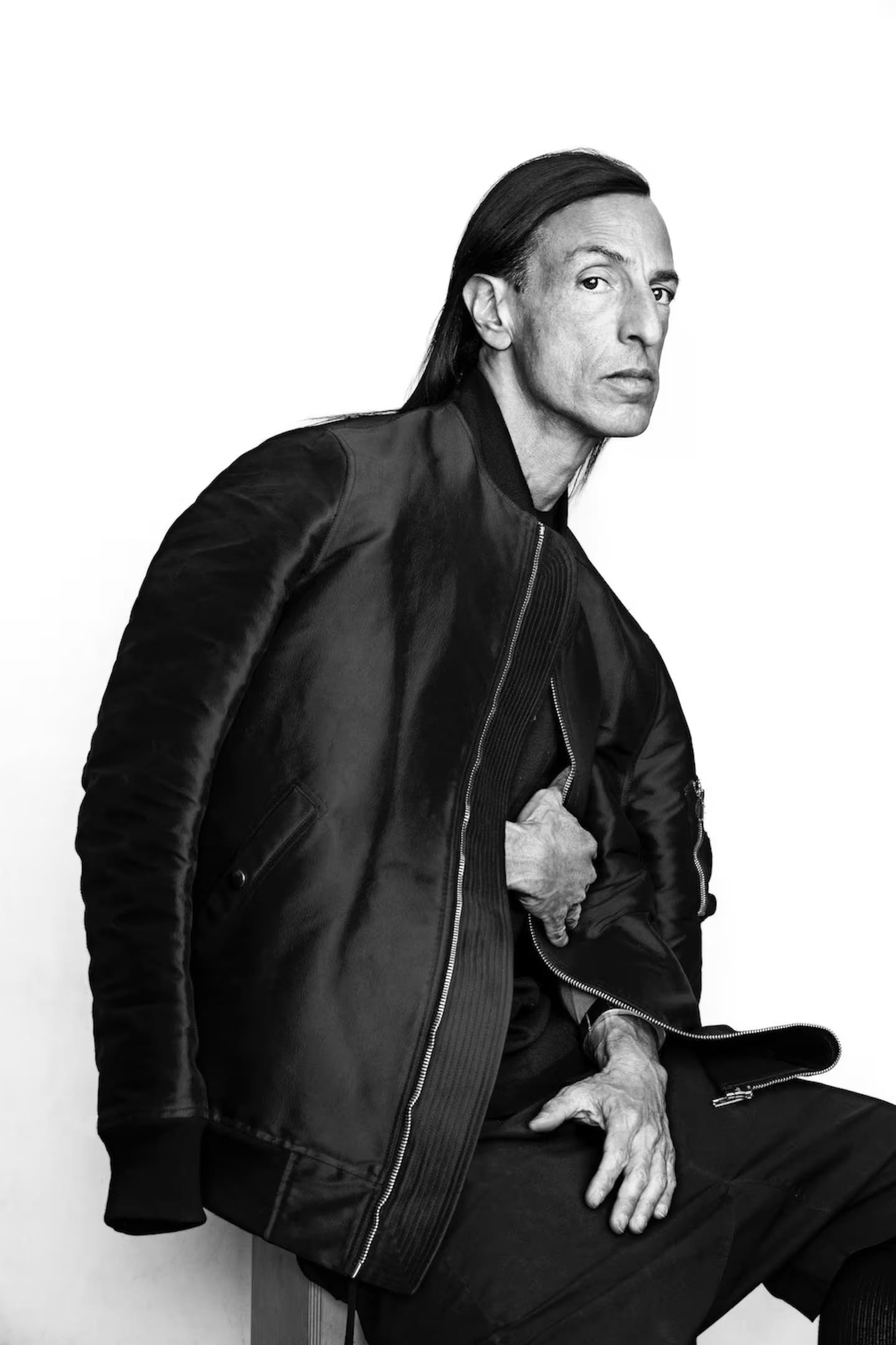 Rick Owens photographed in black and white against a white wall, seated in a black leather bomber jacket.