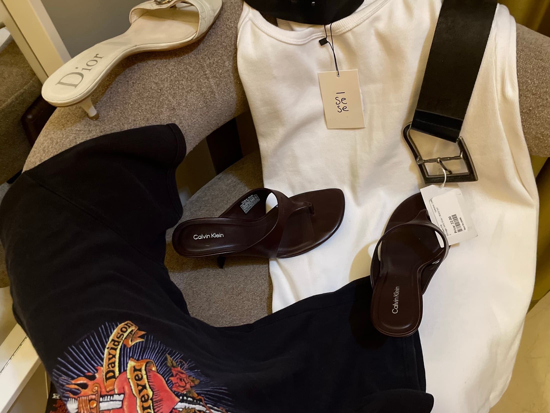 A flat lay on a grey chair: a white tee with its tag still on, a leather belt, two pairs of Calvin Klein sandals and a Dior mule