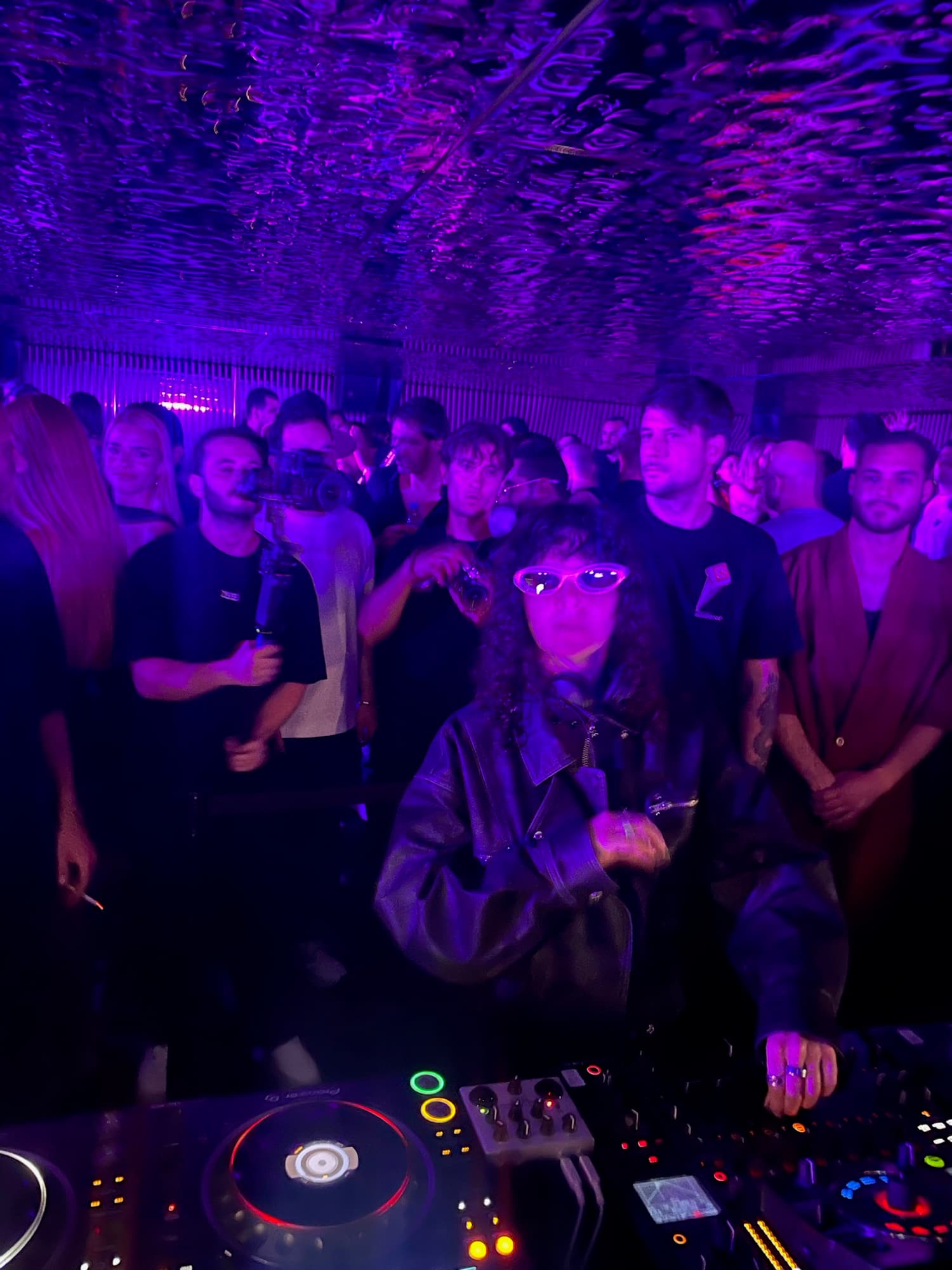 A DJ in sunglasses behind the decks in a crowded room under purple light
