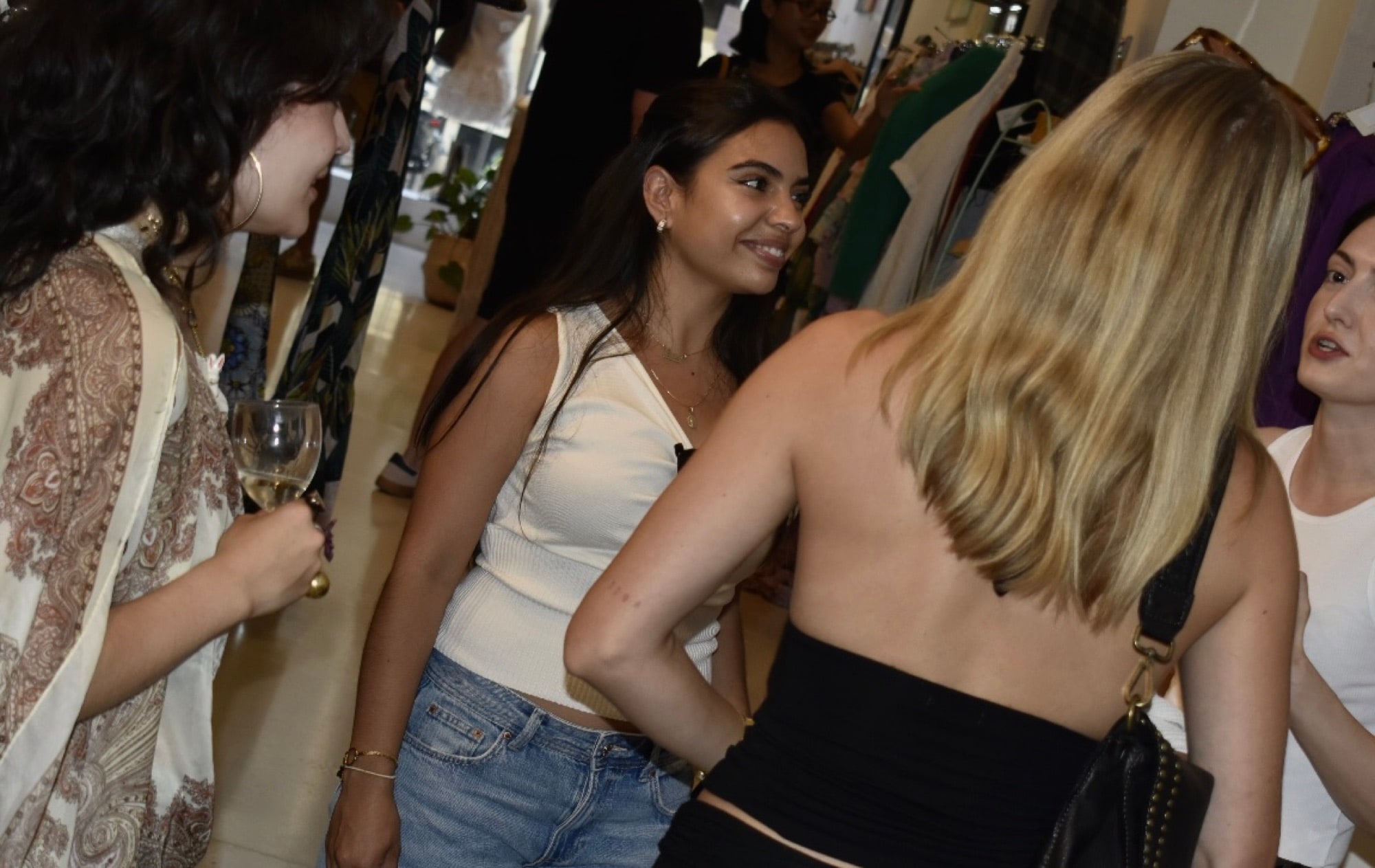 Guests talking between clothing rails at an evening event, one holding a glass of white wine, another in a backless black top.
