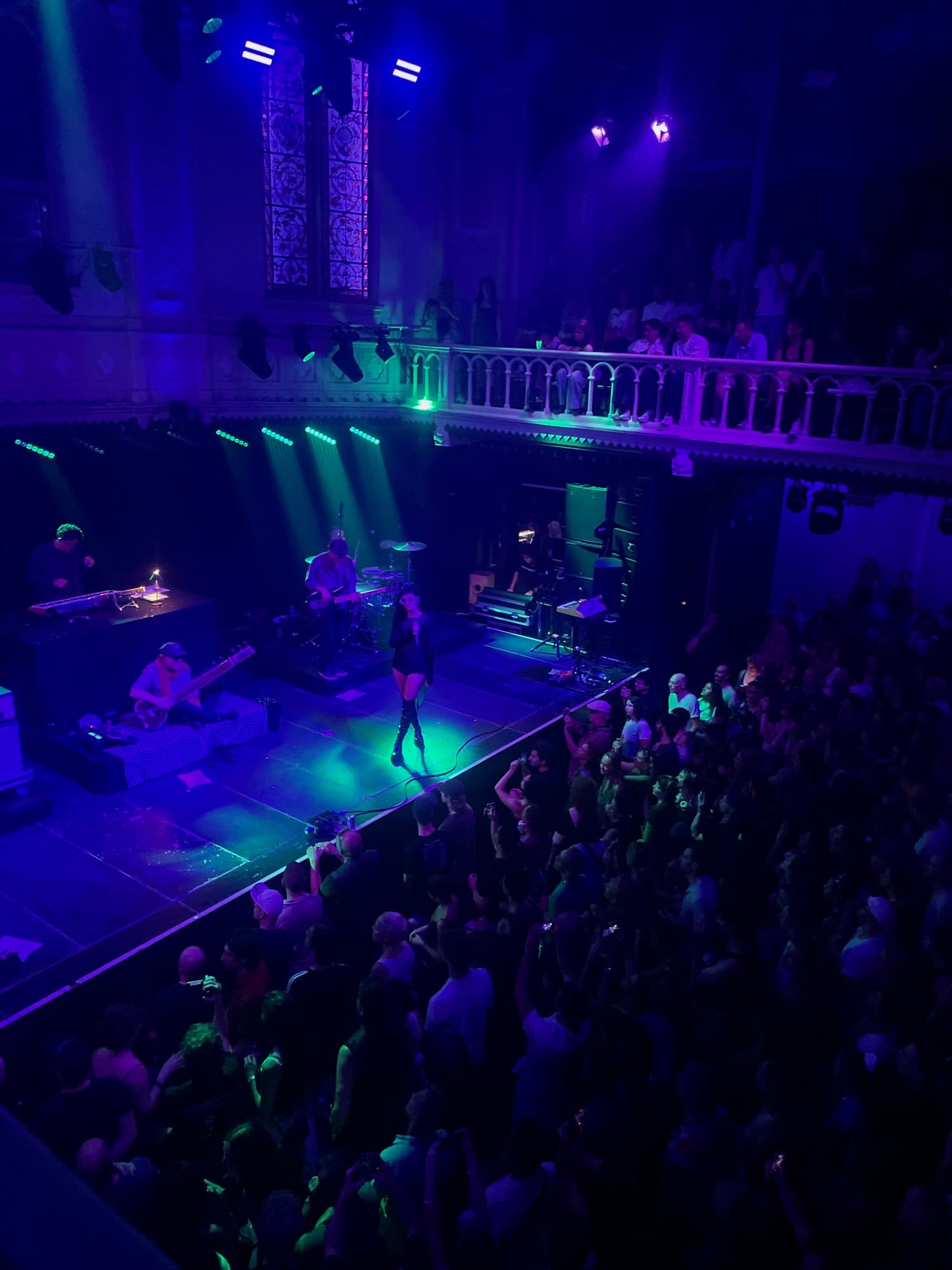 A packed venue seen from the balcony, the stage lit blue