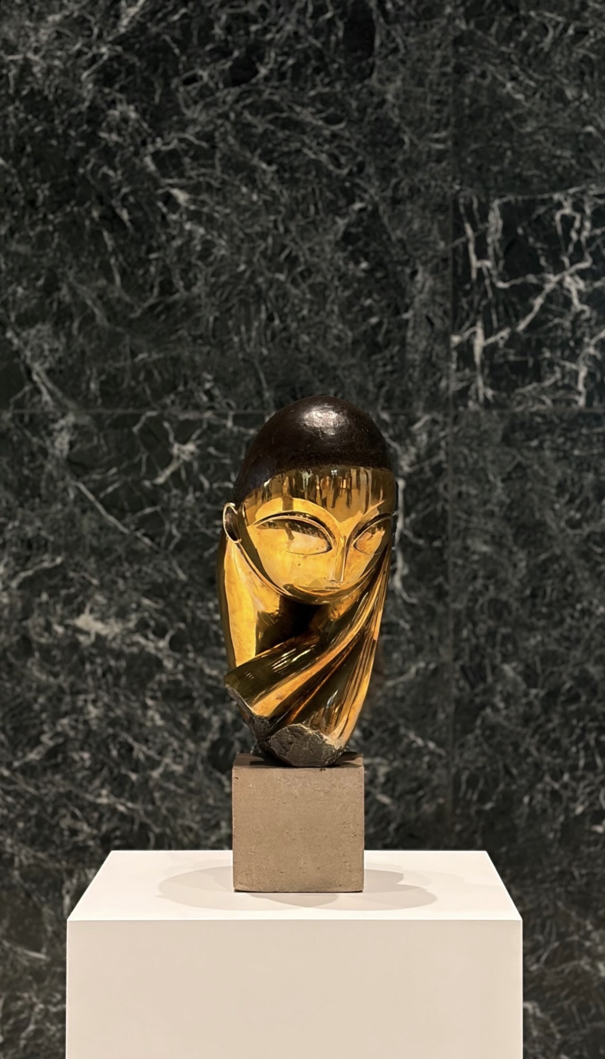 A polished bronze head by Constantin Brancusi on a stone plinth, against dark green marble.