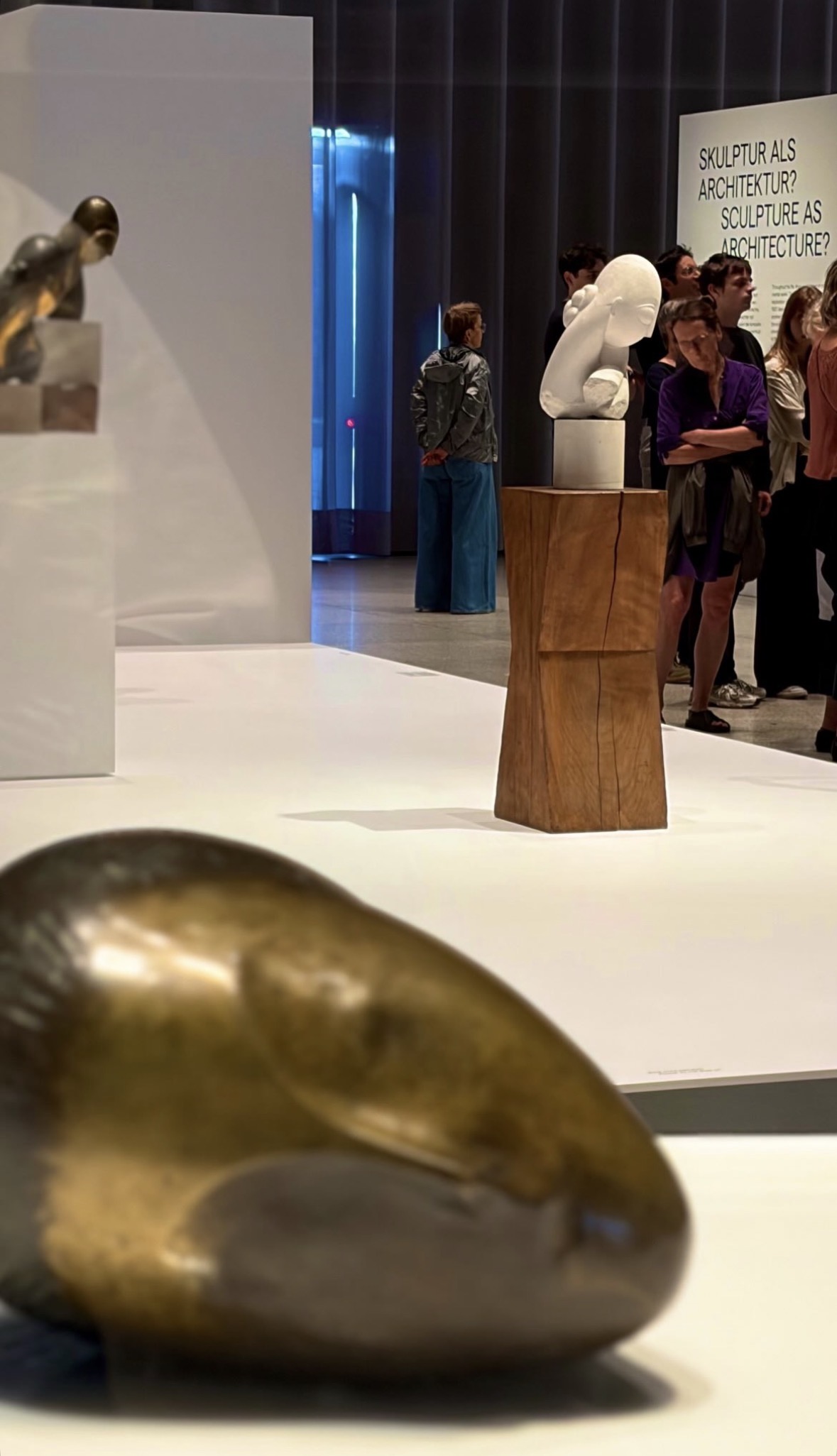 Visitors in a Brancusi exhibition, a white marble head on a carved wooden plinth, beneath wall text reading 'Sculpture as architecture?'
